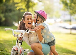Photo of mom and toddler daughter in pink helmet near kid's bicycle with training wheels.